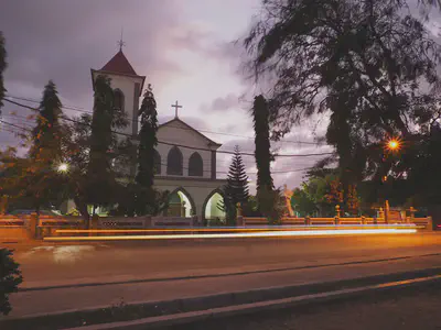 Motael church at dusk with a steeple with a weather vane at the top on the left of the main building with a cross on the roof. A long exposure has captured the light of a motorcycle as a yellow line on the road in front of the churhc.