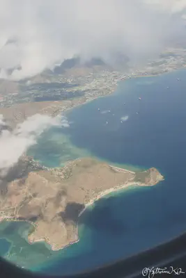 An aerial view of Dili, Timor-Leste. The Cristo Rei statue is on the point with beaches extending from either side. The shallow reefs are visible disappearing into the deep, blue ocean with the city extending up and disappearing into the white clouds.