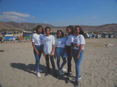 Five young Timorese all wearing the same white &ldquo;Viva Timor-Leste&rdquo; t-shirt, jeans, and sneakers standing on the beach in Tasi Tolu waiting for the 20th anniversary of the referenum. There are small vans selling food lined up behind on the beach with small, brown mountains in the background framed with a blue sky.